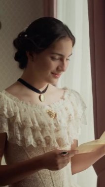 Dikey orta boy, zarif, Viktorya döneminden kalma dantel elbiseli bir kadın elinde aşk mektubuyla pencerede duruyor, okuyor ve göğsüne yakın tutuyor ve gülümsüyor.