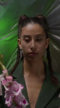 Saçları örgülü, elinde pembe çiçekler tutan, yeşil ışık aksanıyla dokulu gri arkaplanda duran, dikey orta boy bir çift ırklı kadın portresi.