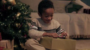 Neşeli Afrikalı Amerikalı çocuk oturma odasında oturmuş Noel hediyesi kutusunu açıyor, baston şeker alıyor ve şapka örüyor.