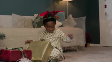 Pijama giyen mutlu Afrikalı Amerikalı çocuk oturma odasında yerde otururken Noel hediyesi kutusunu açıyor.