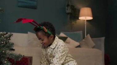 Pijama ve geyik başlığı takan neşeli siyah çocuk Noel hediyelerini açmak için koşuşturuyor, Noel ağacının altında kutuları sallıyor.