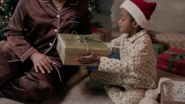 Noel Baba şapkalı pijama giyen Afro-Amerikalı çocuk sevgi dolu annesine Noel hediyesi kutusu veriyor.