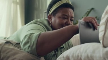 Üst düzey Afro-Amerikan kadının oturma odasında koltukta dinlenip laptopu açması ve internetten bilgi araması.