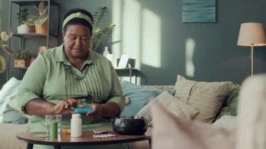 Modern son sınıf siyahi kadının rahat oturma odasında oturmuş diyet takviyeleri alırken orta boy bir fotoğrafı.