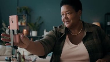 Kıdemli Siyahi Kadın Etkilenen kişi masada oturmuş akıllı telefon kamerasını açıyor ve takipçilerini selamlıyor.