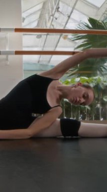 Dikey çekimde siyah tayt giymiş beyaz bir balerin modern bale stüdyosunda yerde esneme hareketleri sergiliyor.