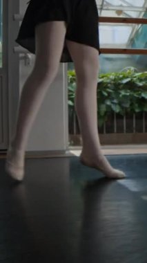 Gündüz antrenman yaparken geniş, parlak stüdyoda siyah tayt giyen, bale antrenmanı yapan genç beyaz bir kızın dikey görüntüsü.