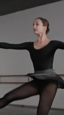 Geniş bale sınıfında zarif bale hareketleri yapan siyah tayt ve tayt giymiş güzel beyaz bir kadının dikey orta pozu.