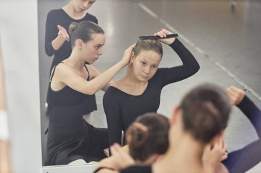 Yansımada yakalanan genç bayan bale dansçısı saçını dikkatlice tararken arkadaşı da stüdyo gösterisine hazırlanırken saç bakımına yardım ediyor.