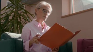 Orta yaşlı, kendine güvenen, kısa gri saçlı ve gözlüklü olgun bir iş kadınını modern güneşli ofiste otururken çekmiş. Turuncu klasördeki belgeleri okumaya ve küçük not defterine not almaya odaklanmış.