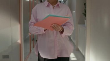 Zarif pembe gömlekli ve gözlüklü beyaz iş kadınının pastel renklerde şık ofis koridorlarında yürürken doküman imzalarken görüntüsünü kaldır ve üretkenliğe odaklan.