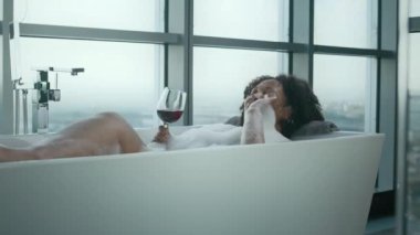 Şehvetli siyahi kadın, yüksek tavan pencerelerinden şehre bakan rahat ve lüks bir atmosferde köpüklerle banyo yaparken kırmızı şarap içiyor.