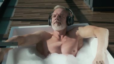 Rahatlamış gri saçlı beyaz adam, panoramik pencerelerden gelen güneş ışığı altında beyaz serbest küvette yatarken kablosuz kulaklıkla müzik dinliyor.