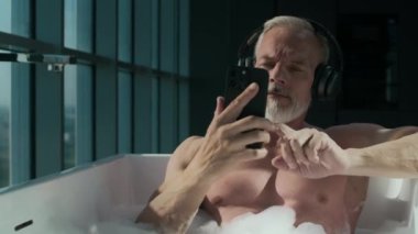 Kablosuz kulaklıklı yakışıklı beyaz adam sabunlu suyla dolu küvette dinlenirken akıllı telefon kullanıyor.