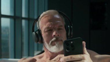 Yakışıklı, gri sakallı adam. Kablosuz kulaklıkla müzik dinliyor ve güneş ışığında küvette dinlenirken akıllı telefondan kayıyor. Yerden tavana manzaraya bakıyor.