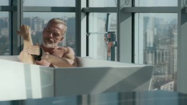 Beyaz saçlı ve sakallı yakışıklı beyaz adam banyo süngeri ile yıkanırken sabunlu suyla dolu küvette oturuyor, yerden tavana doğru dizilmiş.