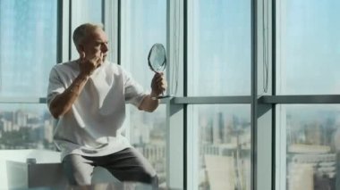 Kafkasyalı bir adamın beyaz küvetin üzerinde otururken, yerden tavana kadar uzanan pencerelere karşı otururken, güneş ışığı yüz kremi sürerken ve aynada kendine bakarken orta boy fotoğrafı.