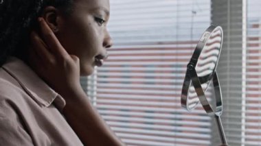 Güzel siyahi bir kadının, tuvalette cilt bakımı için hazırlanırken yuvarlak aynada yüzüne dikkatle baktığı orta boy yakın çekim.