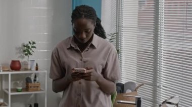 Genç siyahi bir kadının rahat bir pijama giyip rahat, parlak bir banyo ortamında durduğu ve akıllı telefonuyla mesajlaştığı orta boy bir fotoğraf.