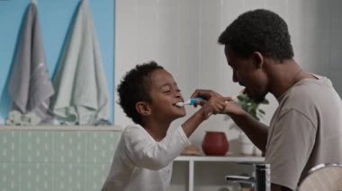 Siyahi baba ve küçük oğlunun sabahları şık, parlak banyoda birbirlerinin dişlerini fırçalarken eğlendikleri orta boy bir fotoğraf.