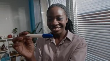 Neşeli genç siyahi kadın ellerindeki pozitif hamilelik testine bakıp gülümsüyor. Parlak, modern banyoda dururken mutluluk ve şaşkınlıkla tepki veriyor.