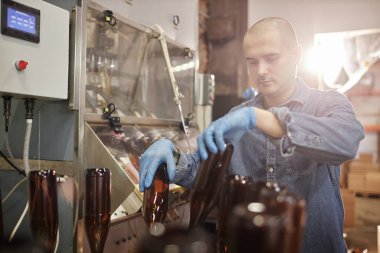 Medium shot of concentrated male production operator in protective gloves carefully sterilizing glass bottles at craft cider or beer factory, copy space clipart