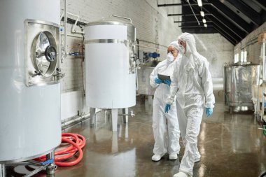 Full length shot of professional female supervisor in mask and coverall walking towards stainless tank with liquid while inspecting workshop equipment with colleague at factory clipart