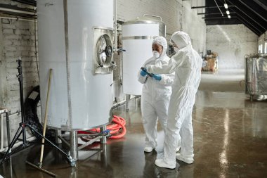 Full length shot of two professional female supervisors in masks and coveralls using tablet computer discussing production process while inspecting tanks in factory workshop clipart