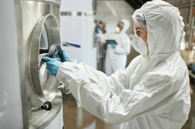 Tehlikeli madde giysisi giyen kadın işlem teknisyenleri ve koruyucu eldivenler, fabrikadaki elma şarabı fermantasyon ekipmanlarını incelerken, sıvı dolu akvaryumun vanasını çeviriyorlar.