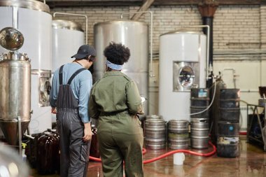 Rear view of diverse team of two cider factory workers inspecting production process on workshop floor with huge fermentation tanks at plant, copy space clipart