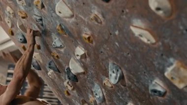 Spor şortu giyen güçlü, orta yaşlı bir sporcunun iç mekan tırmanma merkezinde yapay kayaya tırmanırken ki görüntüsü.