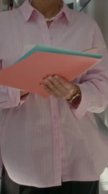 Kendine güvenen beyaz iş kadınının zarif pembe tişört ve gözlük takıp, şık ofis koridorlarında yürürken belge imzalarken, üretkenliğe odaklandığı dikey pozlar.