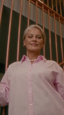 Düşük açılı, orta ölçekli, açık pembe gömlekli, kısa gri saçlı, modern, gösterişli ofis merdivenlerinde kameraya poz veren olgun beyaz iş kadını portresi.