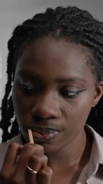 Açık mavi duvarlı ve beyaz kapılı minimalist banyoda dişlerini fırçalayan genç siyahi kadının dikey yakın çekimi.