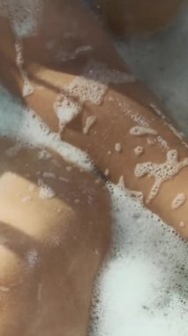Küvette sıcak su ve sabun köpükleriyle banyo süngeri kullanan kimliği belirsiz bir kadının dikey görüntüsü. Evde sabah rutini sırasında kendi başının çaresine bakıyor.