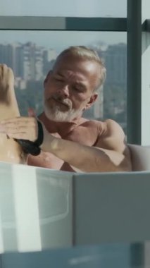 Beyaz saçlı ve sakallı yakışıklı beyaz adamın dikey hâli banyo süngeri ile yıkanırken sabunlu suyla dolu küvette oturması, tavandan tavana karşı kurulması.