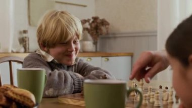 Yeşil bardaklarda sıcak içeceklerle masada oturan iki genç çocuk büyük anne ve babalarıyla kırsal evlerde satranç oynuyorlar.