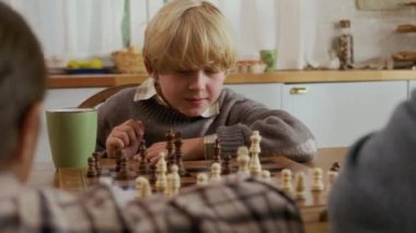 İçerde baba veya büyükbabayla satranç oynayan odaklanmış bir çocuk sandık dolusu satranç tahtası ve ahşap masada duran yeşil kupayla bir sonraki hamlesini düşünceli bir şekilde planlıyor.