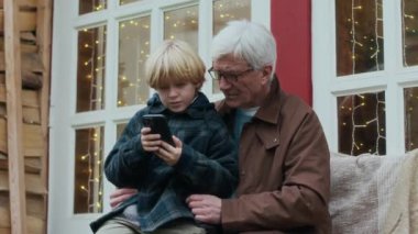 Orta boy beyaz bir büyükbaba ve torunu, sonbaharda ışıldayan sıcak ışıklarla dekore edilmiş verandada akıllı telefonlara bakarken kucağında oturuyorlar.