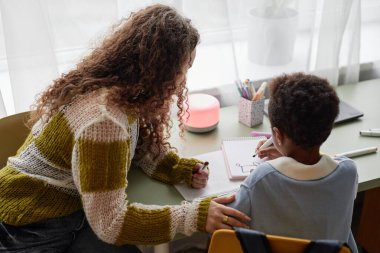 Siyahi bir çocuk ve destekleyici bir annenin renkli keçeli kalemlerle çizim yaparken çocuk odasında masasında yan yana otururken çekilmiş gizli bir fotoğrafı.