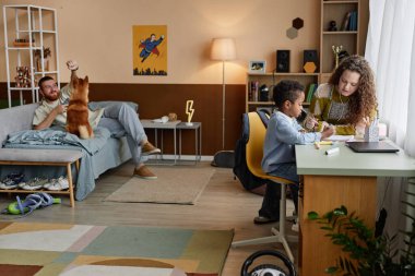 Geniş açılı çekimde, küçük siyah çocuk, annesiyle birlikte çocuk odasında resim çizerken, baba arka planda köpekle oynarken, sıradan bir ev hayatı sahnesi, kopyalama alanı.