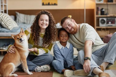 Küçük siyah çocuğun portresi, neşeli üvey ailesi ve şirin Shiba Inu köpeğiyle çocuk odasında oturmuş kameraya gülümsüyor, mutlu aile kavramı.