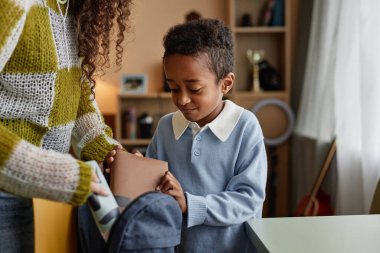 Siyahi çocuğun okul çantasına kitap ve kırtasiye malzemesi koyarken tanınmayan üvey annenin, evlat edinilen çocuğun okula hazırlanmasına yardım ettiği orta boy bir fotoğraf.