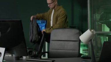 Genç beyaz erkek programcının gündelik kıyafet ve gözlük taktığı orta boy bir fotoğraf modern çalışma ofisine varıyor, kulaklık takıyor, masa lambasını açıyor ve geceleri bilgisayarda çalışmaya başlıyor.
