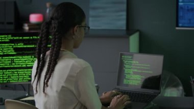 Gözlüklü konsantre genç kadın çift ırklı programcının arka plan görüntüsü. Günlük kıyafet giyiyor. Dizüstü bilgisayar programında çalışıyor ve geceleri modern bilgisayar ofisinde bilgisayar başında çalışıyor.