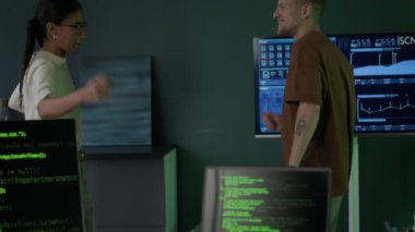 Genç kadın melez programcı ve beyaz erkek arkadaşının dans edip ön planda bilgisayar ve dizüstü bilgisayar ekranlarında program kodlarıyla eğlenirken yavaş yavaş aşağı eğilmeleri.