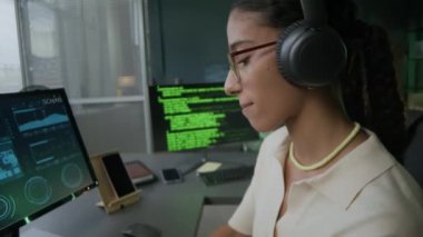 Relaxed young female biracial programmer in eyeglasses and casualwear enjoying listening to music in headphones while working on PC in modern programmers office at night