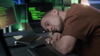 Yorgun genç beyaz erkek programcı masada uyuyor ve telefon tebligatı ile uyanıyor, gözlerini gece modern ofis arka planında çalışan program kodlarıyla ovalıyor.