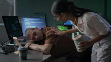 Genç kadın melez programcı, yorgun beyaz erkek arkadaşını uykusundan uyandırıp gece vakti modern programcıların ofisinde program kodlaması üzerinde çalışmaya devam ediyor.