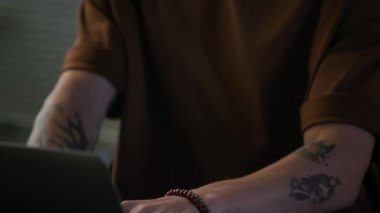 Genç, konsantre olmuş beyaz erkek programcının dövmeli kollarıyla dizüstü bilgisayarda yazarken gece ofisindeki masada otururken görüntüsünü kaldır.